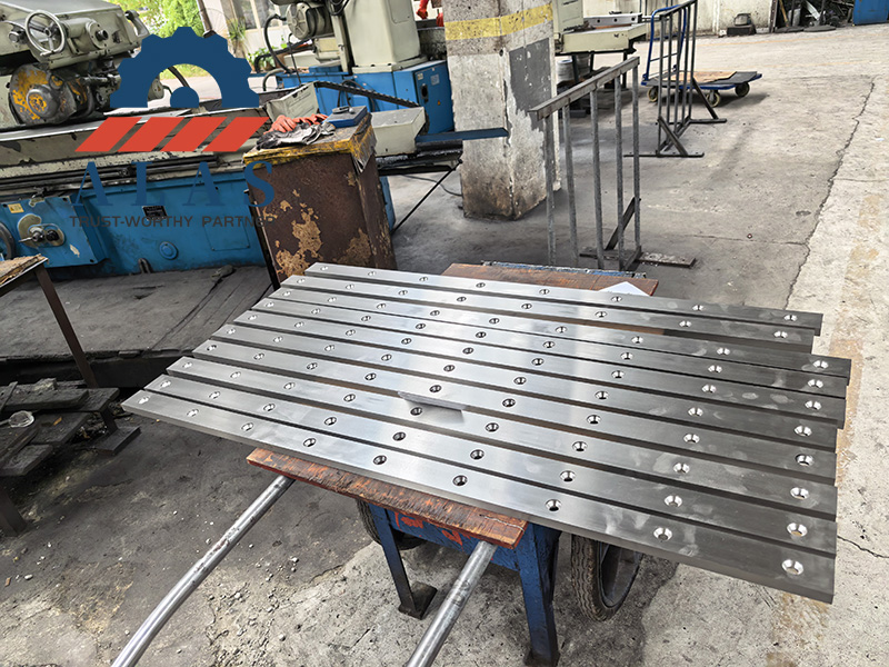 A batch of precision-ground guillotine shear blades with counterbored mounting holes stacked in the ALAS manufacturing workshop